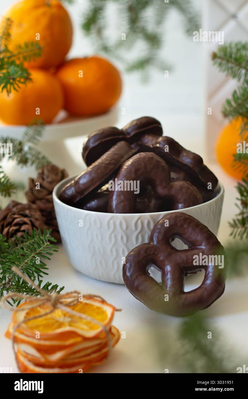 Köstliche Brezeln mit Schokoladenüberzug stapeln sich in einer weißen Schüssel, umgeben von festlichem Grün und frischen Orangen. Diese reizvolle Szene fängt ein W Stockfoto