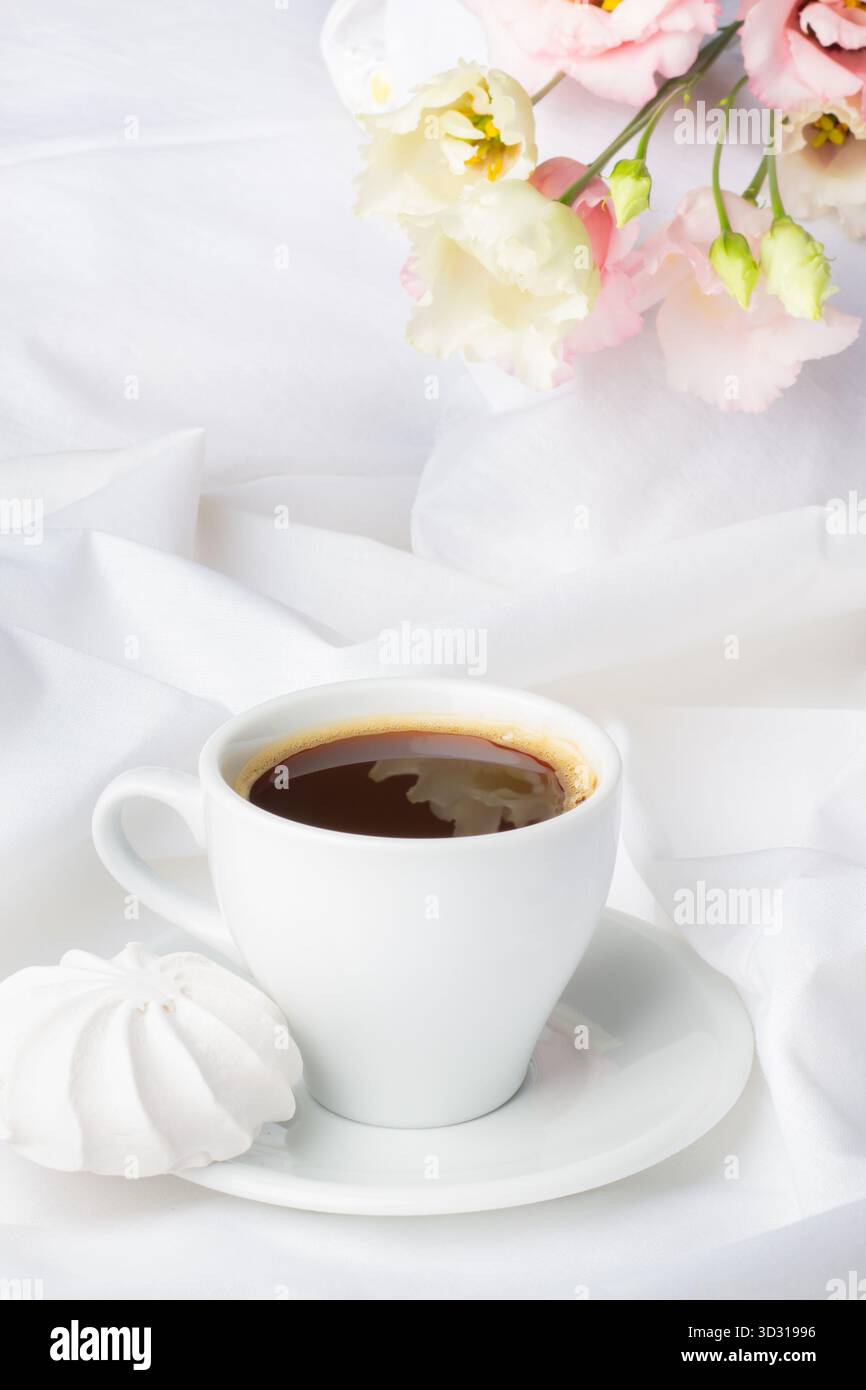 Eine weiße Kaffeetasse mit dunklem Kaffee steht neben einem weichen Meringue-Keks, umgeben von frischen Pastellblüten auf seidenweißem Stoff, wodurch ein s entsteht Stockfoto