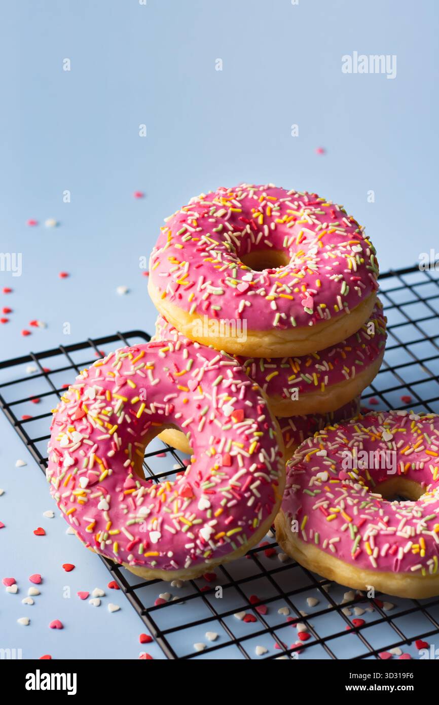 Vier rosafarbene Donuts mit bunten Streuseln sind auf einem Kühlregal aus Metall gestapelt. Bunte Streusel streuen sich herum und sorgen so für Spaß Stockfoto