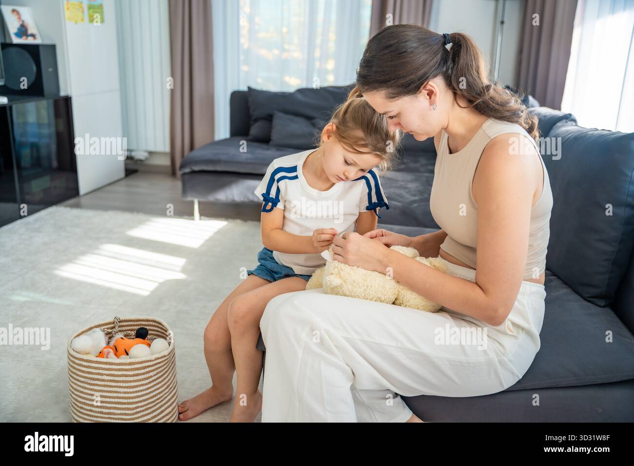 Mutter und Tochter reparieren zusammen einen Teddybären, während sie zuhause sitzen. Gemeinsame Momente der Liebe und Bindung durch alltägliche Rituale und Stockfoto