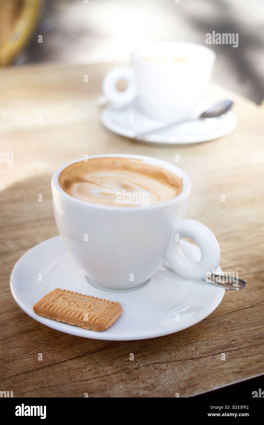 Nahaufnahme eines Cappuccinos, serviert in einer weißen Keramikschale mit Illy-Logo auf Holztisch mit einem Keks und einer anderen Tasse, die im Hintergrund verwischt ist palma mallorca Stockfoto