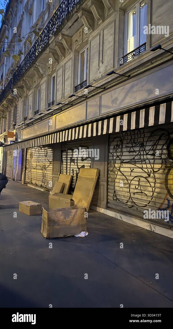 Pappkartons wurden an einer mit Graffiti bedeckten Wand in einer städtischen Straße entsorgt. Die geschlossene Ladenfront mit gestreiften Markisen unterstreicht die ruhige, frühe e Stockfoto