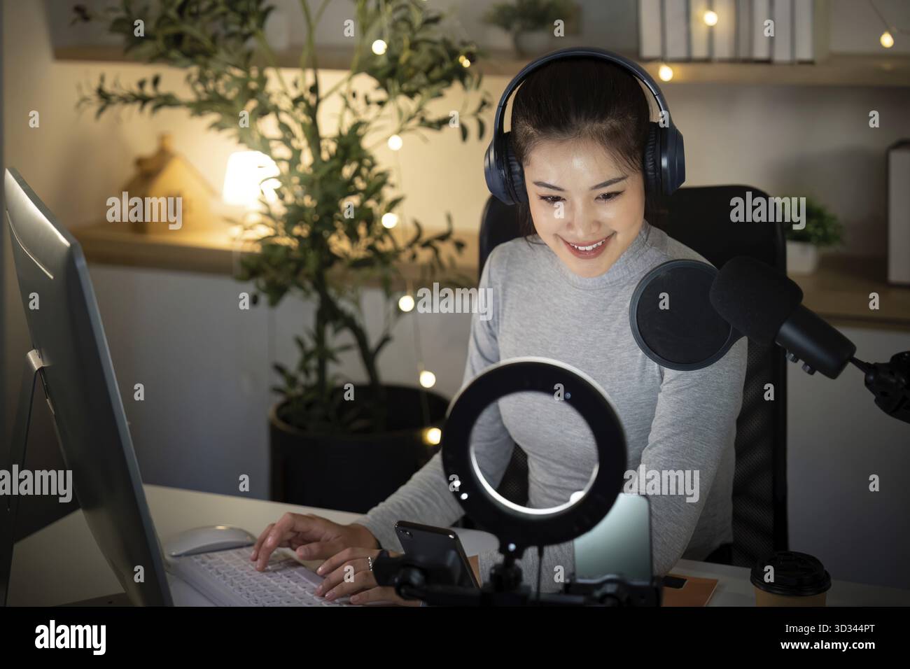 Eine junge Frau mit Kopfhörern sitzt an ihrem Schreibtisch und lächelt, während sie sich auf ihre Podcast-Aufnahme vorbereitet. So entsteht eine warme und einladende Atmosphäre Stockfoto