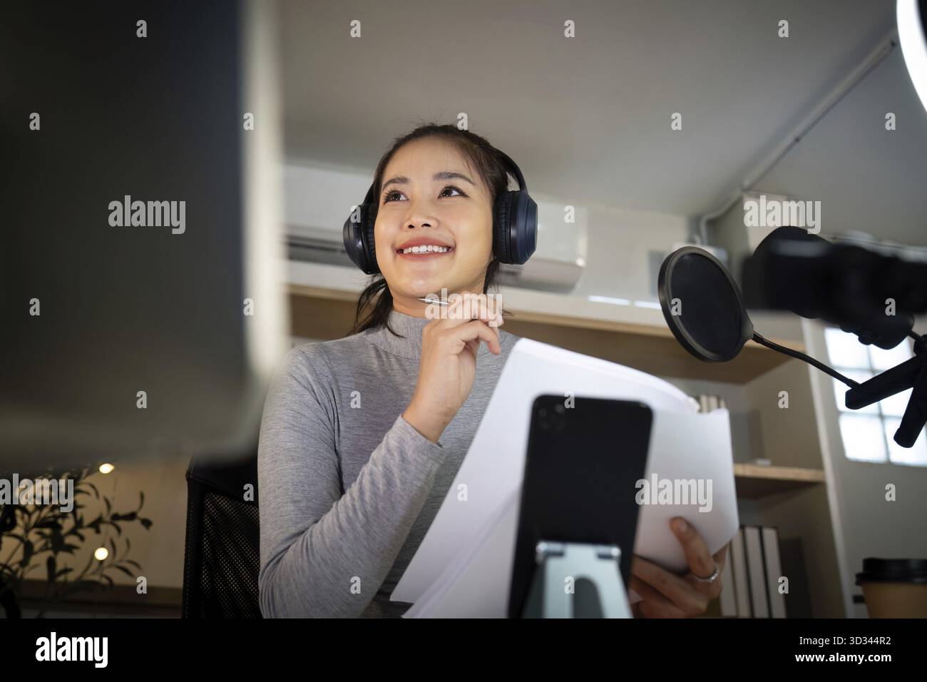 Eine junge Frau lächelt, während sie Notizen hält und einen Moment der Inspiration festnimmt, während sie ihren Podcast in einem hellen Studio aufnimmt Stockfoto