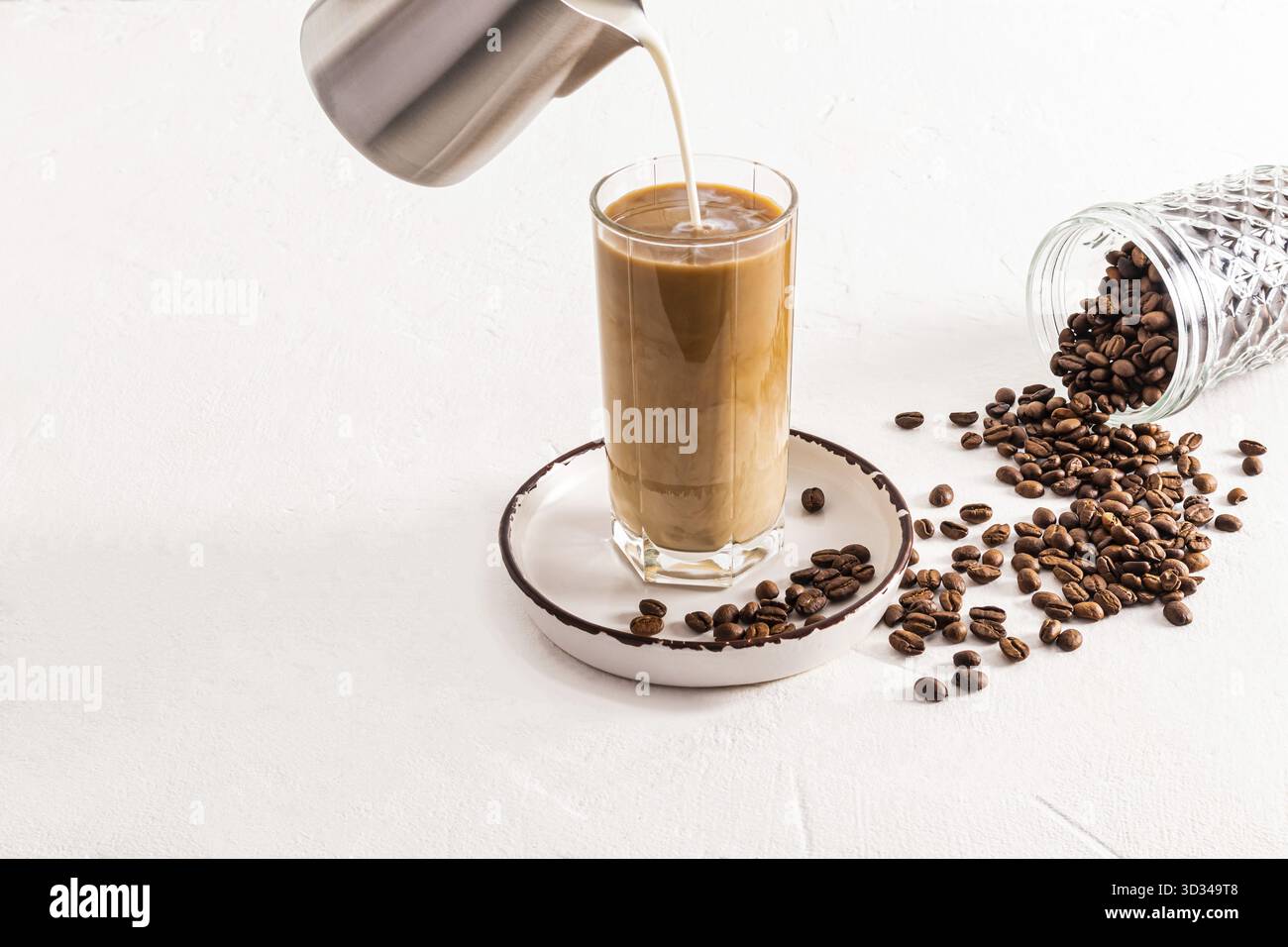 Ein Milchstrahl aus einer Metallkanne wird in ein hohes Glas eiskalten Kaffees gegossen. Dosen zur Aufbewahrung von Kaffee. Zubereitung eines köstlichen Getränks Stockfoto