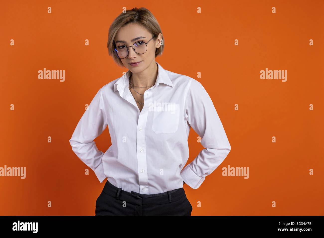 Ehrgeizige Frau, die ein weißes Hemd im offiziellen Stil trägt und mit Händen auf den Hüften auf orangefarbenem Hintergrund steht Stockfoto