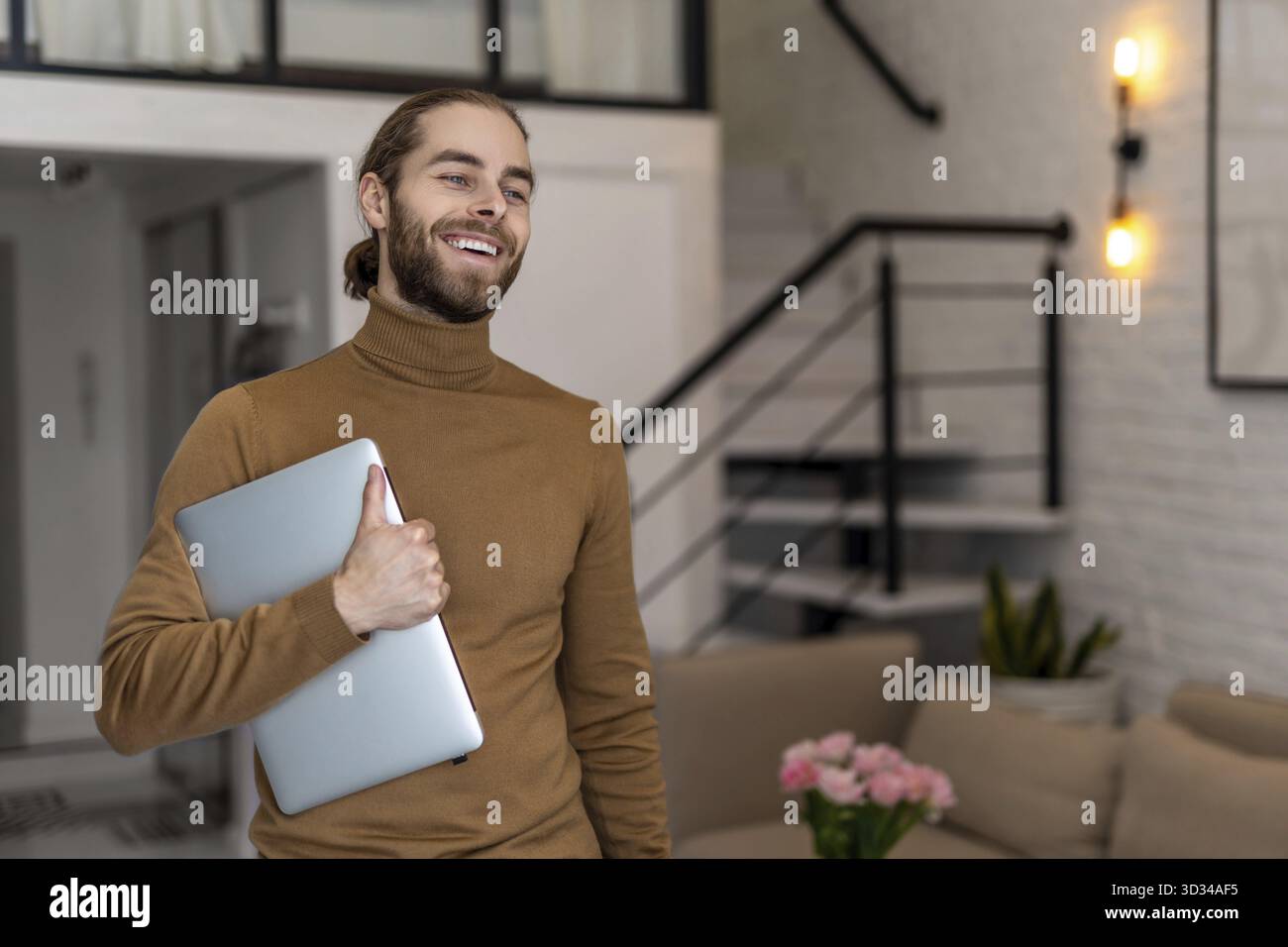 Lächelnder attraktiver Mann mit Bart, der legere Kleidung trägt und zu Hause mit gefaltetem Laptop steht Stockfoto
