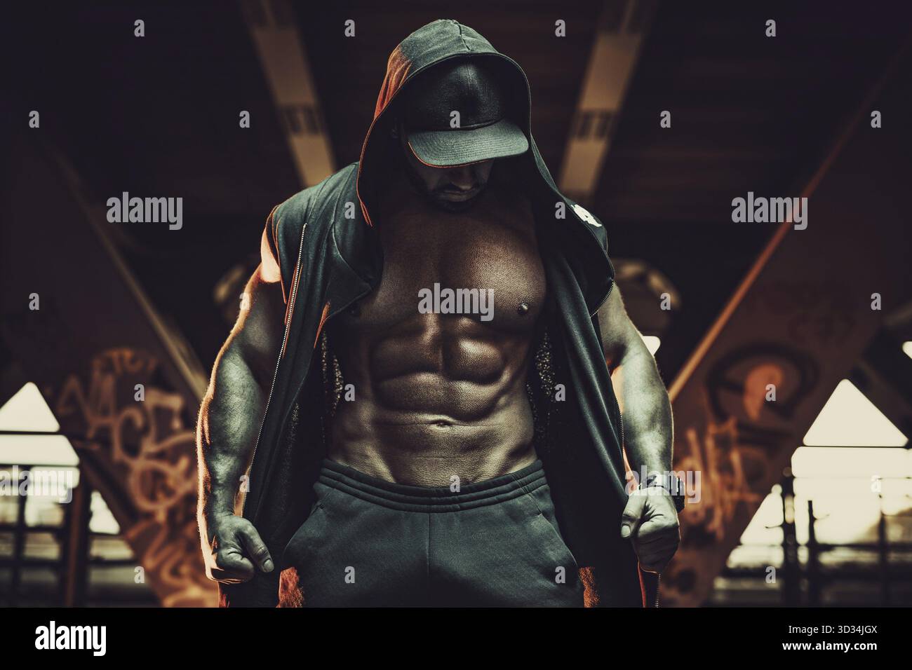 Junger starker Mann Bodybuilder mit ungezwungener Kleidung und Kapuze im urbanen Stadtinneren. Dunkelrote Farben Stockfoto