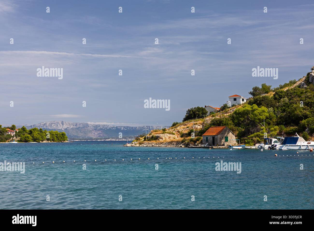 Eine ruhige Küstenlandschaft bietet einen felsigen Hügel mit Bäumen, ein weiß getünchtes Steinhaus mit rotem Dach und kleine Boote, die entlang einer klaren B ankern Stockfoto
