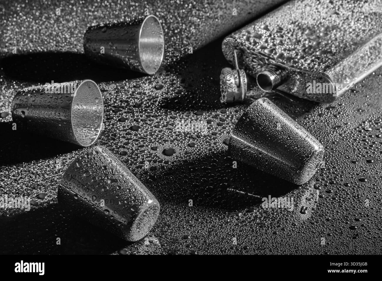Schwarz-weiße Nahaufnahme von Metallkolben und Schussgläsern auf nasser Oberfläche. Wassertropfen und Reflexionen sorgen für ein dramatisches und strukturiertes Bild. Stockfoto