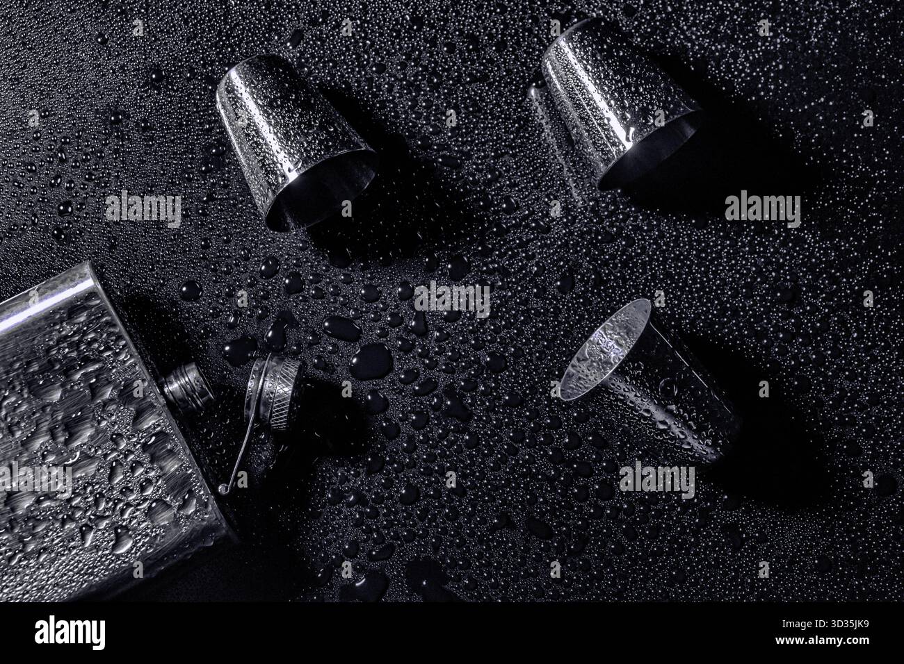 Metallkolben mit offenem Verschluss und drei Shot-Gläsern auf dunkler, nasser Oberfläche. Wassertropfen und Reflexionen sorgen für eine stimmungsvolle, kontrastreiche Darstellung. Stockfoto