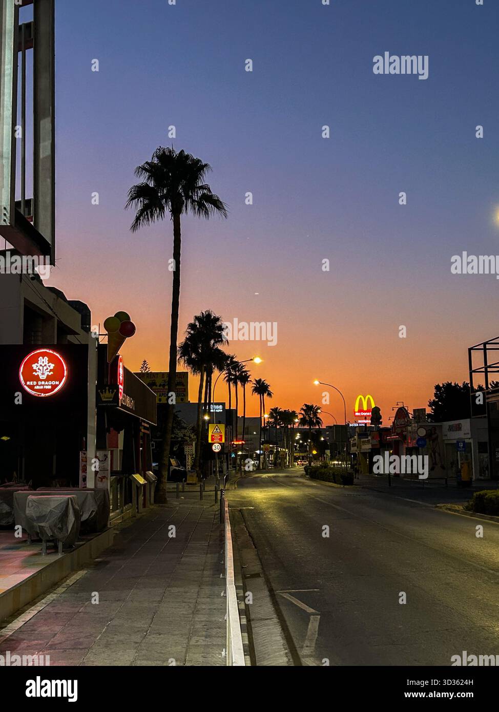 Friedliche Stadtstraße gesäumt von Palmen und leuchtenden Schildern unter einem farbenfrohen Sonnenuntergangshimmel, die die ruhige abendliche urbane Atmosphäre einfangen lässt. - Smartphone-aufgenommenes Stockfoto