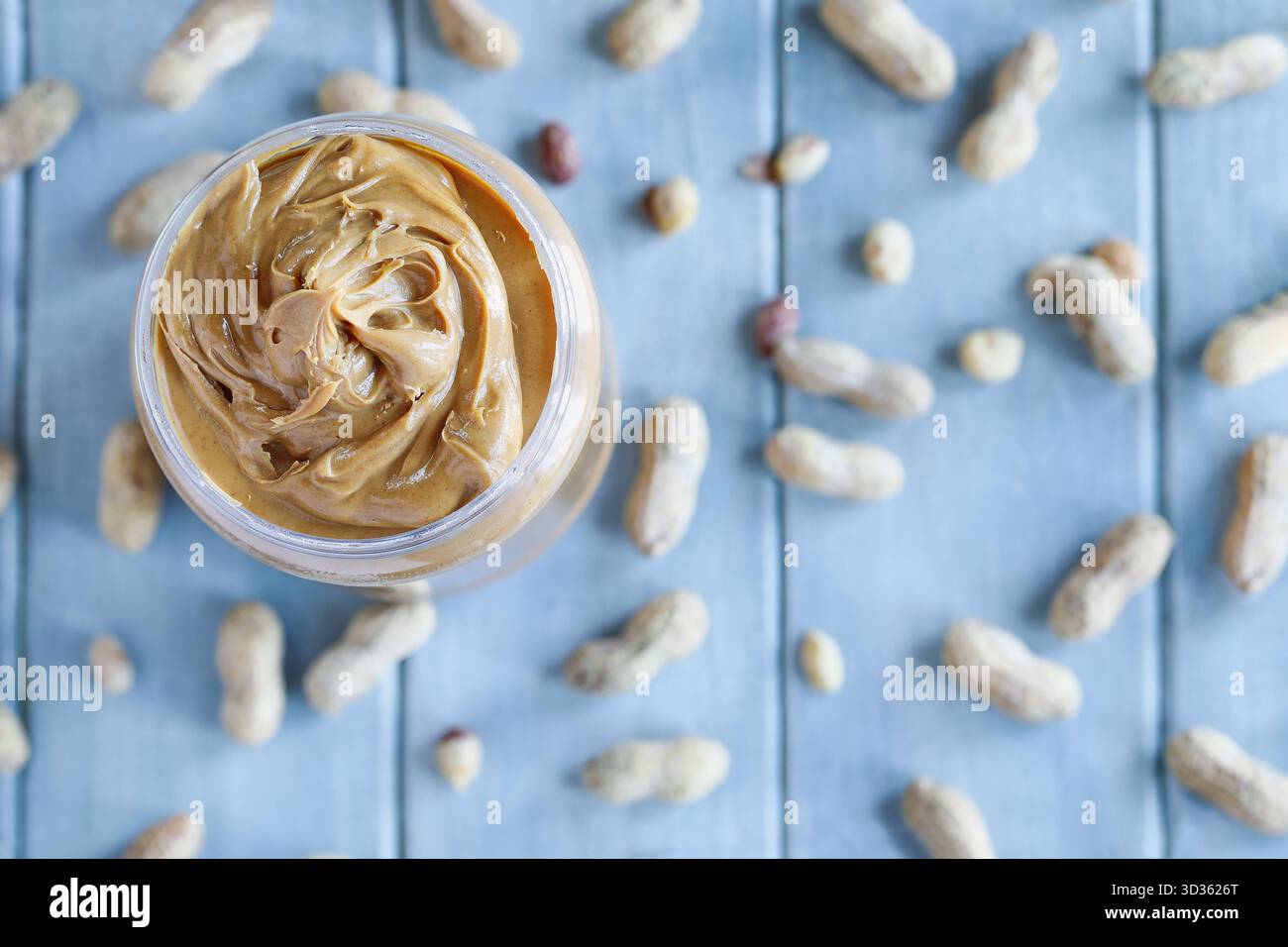 Schwenken Sie cremige Erdnussbutter in einem Plastikbehälter über einem blauen Tisch mit verstreuten Nüssen in der Schale. Selektiver Fokus mit Unschärfe Stockfoto