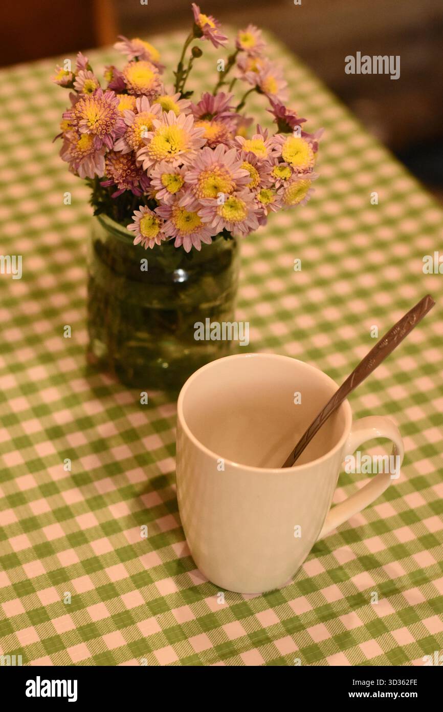 Weiße Tasse mit einem Löffel auf einer grünen karierten Tischdecke, neben einem Glas mit rosa Chrysanthemen. Draufsicht, gemütliches Foto im Heimstil. Stockfoto