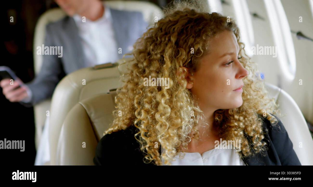 Die lockige Frau im Blazer in der Jet-Kabine, mit cremefarbenen Sitzen, Gürteln, Smartphone, ovalen Fenstern Stockfoto