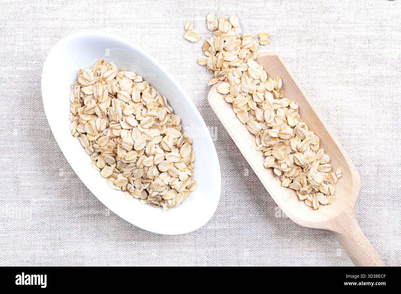 Haferflocken, dickgerollte, altmodische Haferflocken, in weißer Schüssel und Körnerlöffel, auf Leinen. Geschälte, gedämpfte Hafergrieß, zu Flocken gerollt und geröstet. Stockfoto