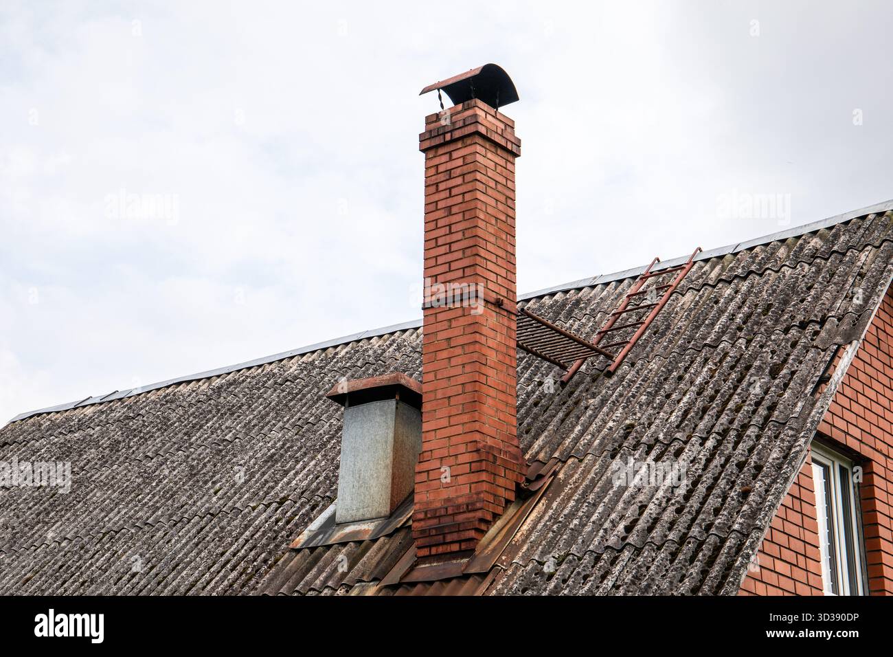 Nahaufnahme eines geneigten Daches mit verwitterten Wellblechen, einem hohen roten Ziegelkamin, einem Metallschacht und einer Zugangsleiter für die Wartung. Stockfoto