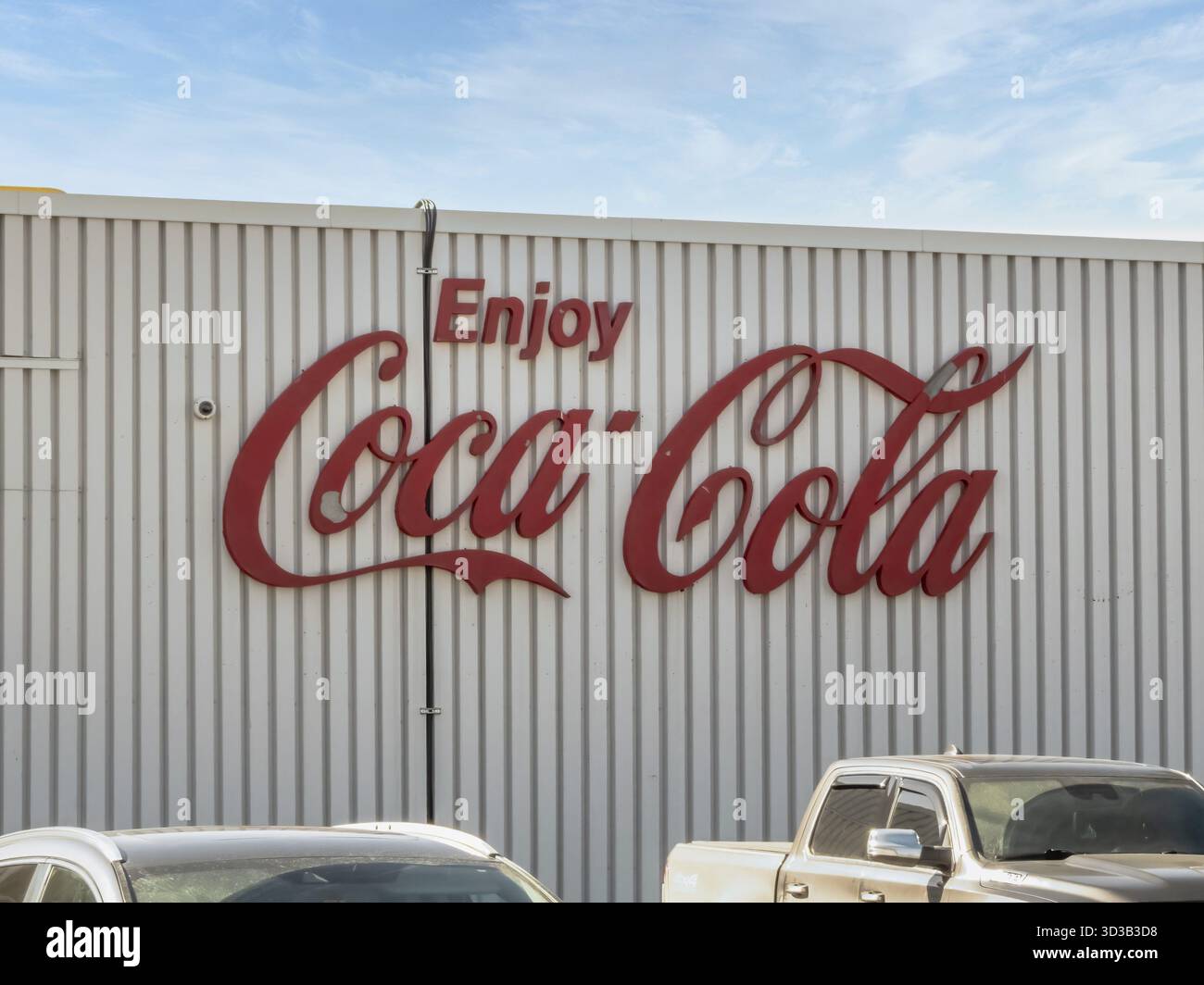 Calgary, Alberta, Kanada. April 2025. Das ikonische rote „Enjoy Coca-Cola“-Logo ziert das gewellte Metallgehäuse eines Gebäudes, das über geparkten Autos sichtbar ist Stockfoto