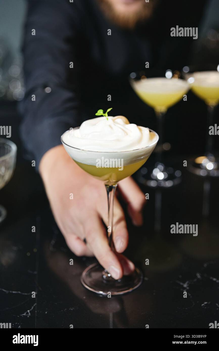 Essen, Nahaufnahme des Barkepers, der einen Zitruscocktail mit dickem Schaum und Kräutergarnitur in einem stilvollen Coupé-Glas präsentiert Stockfoto