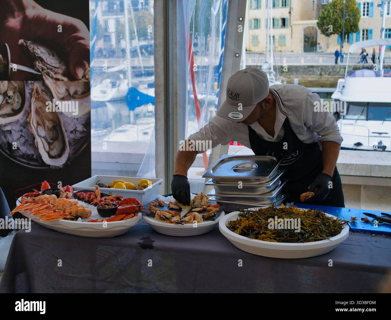 Vannes, Frankreich - September 30 2025: Ein Küchenchef arrangiert bei einem kulinarischen Wettbewerb am Yachthafen in Vannes, Bretagne, Frankreich, eine Platte mit frischen Meeresfrüchten. Im Hintergrund stehen Boote und ein Marktzelt. Stockfoto