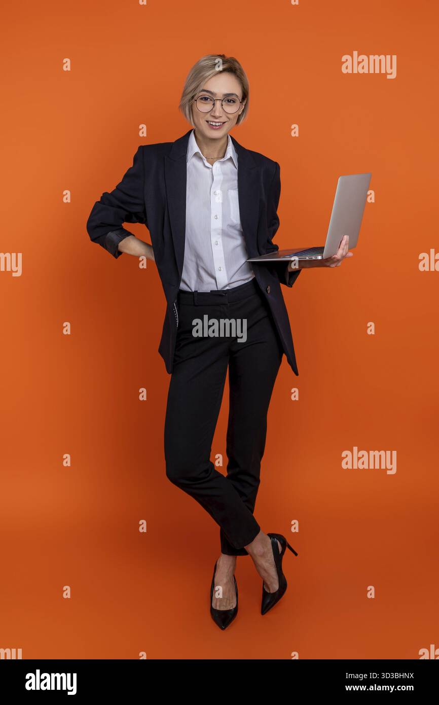 Schöne Frau, die einen schwarzen offiziellen Anzug trägt, der pc isoliert über orangefarbenem Hintergrund hält Stockfoto