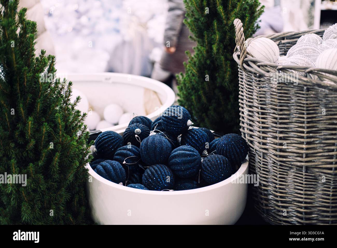 Blauer glitzernder Weihnachtsschmuck in einer weißen Schüssel zwischen künstlichen Kiefern und Korbkörben. Große Weihnachtsdekoration, Schneeballschmuck Stockfoto