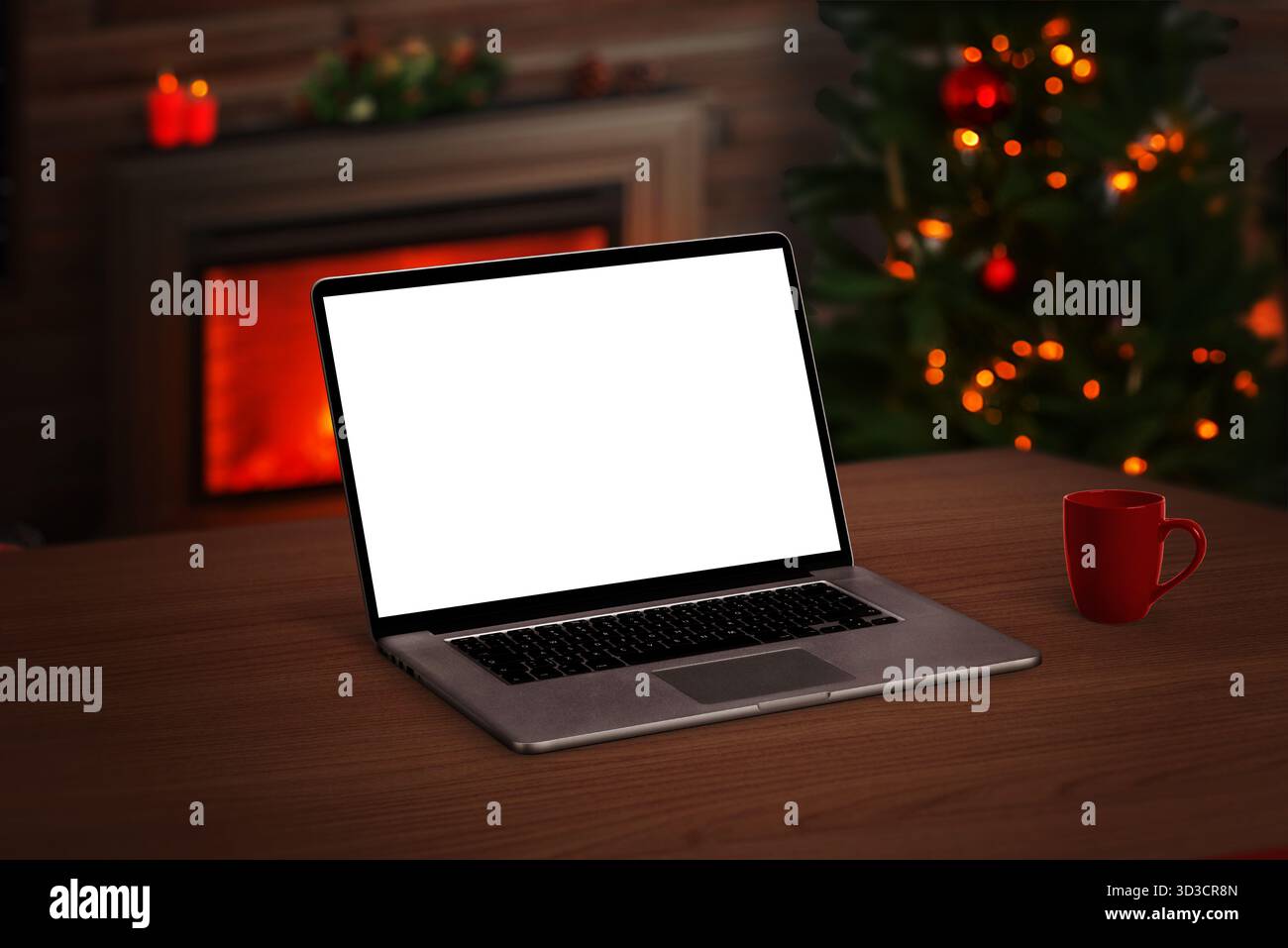 Laptop mit weißem Bildschirm auf Holztisch, gemütlicher Innenraum mit Weihnachtsbaum, Becher und leuchtendem Kamin im Hintergrund Stockfoto