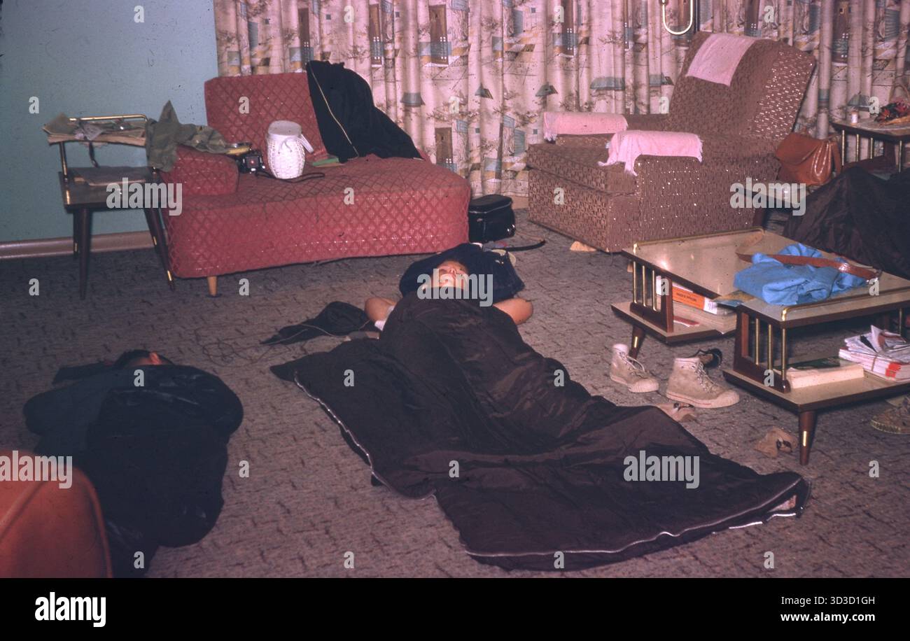 Junge Männer schlafen in Schlafsäcken auf dem Boden eines Wohnzimmers aus der Mitte des Jahrhunderts, das mit einem gesteppten Sofa, einem gewebten Sessel, Tischen mit verjüngten Beinen und malerischen Barttuchvorhängen ausgestattet ist, USA, 1964. Stockfoto