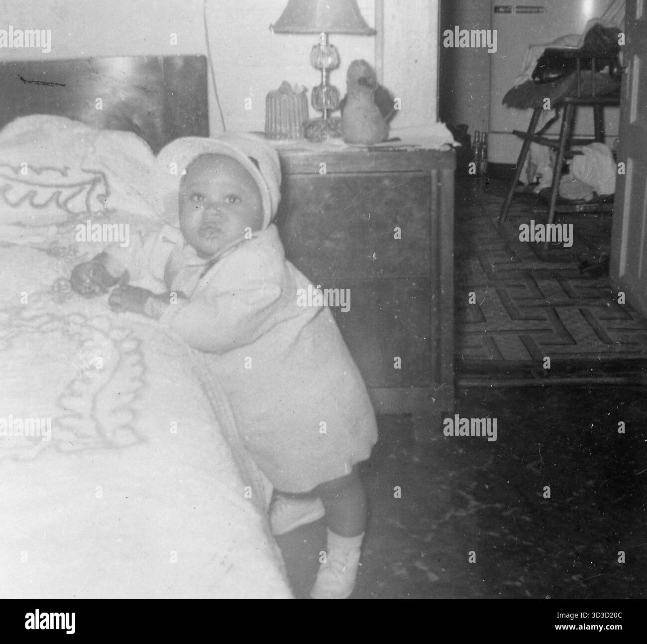 Ein afroamerikanischer Säugling mit Wollmantel, passender Mütze und Schnürschuhen hält sich vor einem mit Chenille bedeckten Bett in einem Haus aus der Mitte des Jahrhunderts, ein Nachttisch mit einer Glaslampe und Stoffspielzeug, Parkettboden und ein hölzerner Hochstuhl mit Limonadenflaschen darunter sind im Hintergrund zu sehen, USA, 1958. Stockfoto