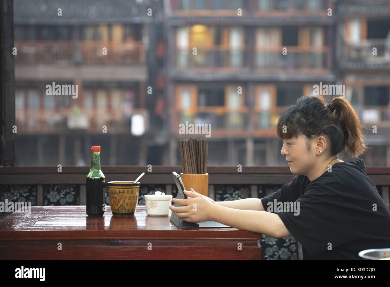 Feng Huang, China - August 2019 : Chinesin sitzt in einem Café-Restaurant und benutzt ihr Tablet, um Clips zu sehen oder zu arbeiten, Fenghuang Old Town Stockfoto