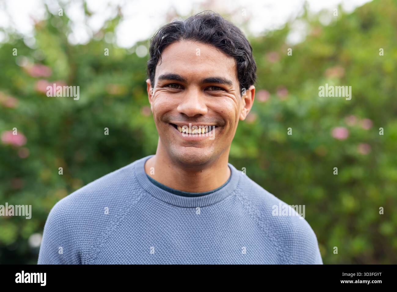 Mann lächelt, während er im Garten steht, in blauem Strickpullover zwischen rosa Blüten und Laub Stockfoto