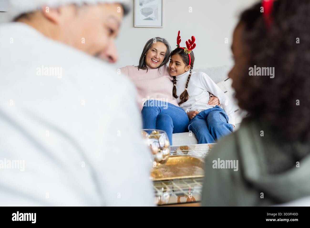 Eine ältere Frau umarmt ein asiatisches Mädchen, das zu Hause auf dem Sofa sitzt, mit Weihnachtsbügeln und einer dekorativen Schüssel Stockfoto