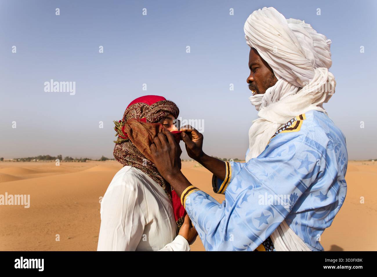 Ein ortsansässiger Mann hilft einer Touristen, ihr traditionelles Kopftuch in den Sahara-Dünen in Merzouga, Marokko, anzupassen und hebt Berbercul hervor Stockfoto