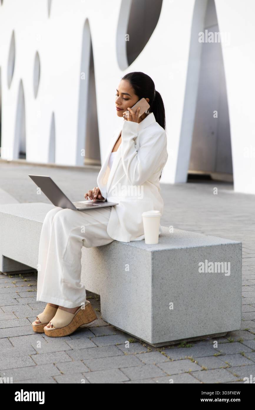 Eine Geschäftsfrau in einem modernen, weißen, urbanen Setting multitaskt nahtlos auf ihrem Laptop, während sie ein Telefongespräch führt, und verkörpert professionelle Vielseitigkeit Stockfoto