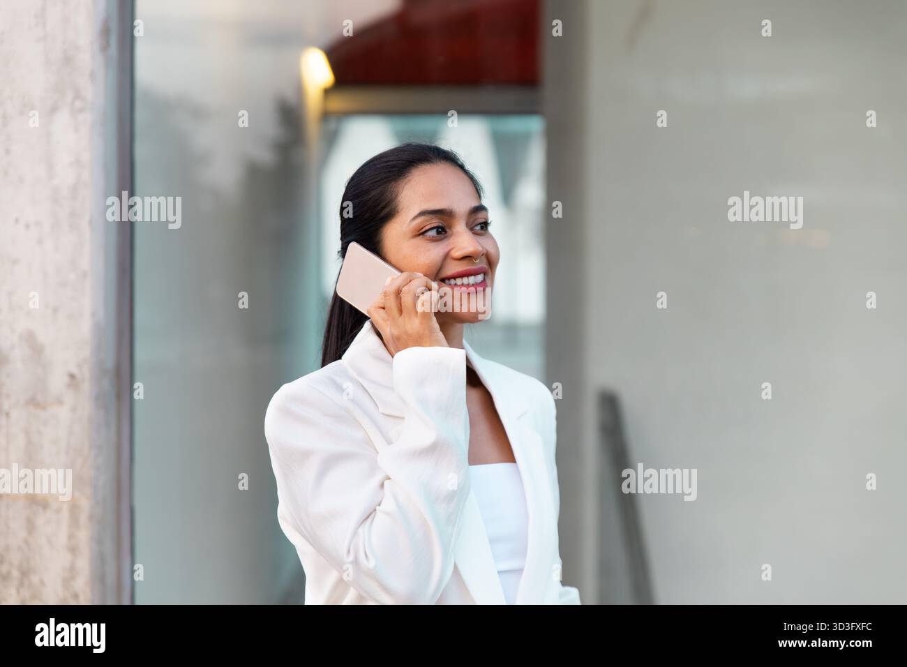 Selbstbewusste Geschäftsfrau in einem eleganten weißen Blazer spricht mit ihrem Smartphone und steht in einer modernen urbanen Umgebung. Ihr entspanntes Auftreten spiegelt das Bekenntnis wider Stockfoto