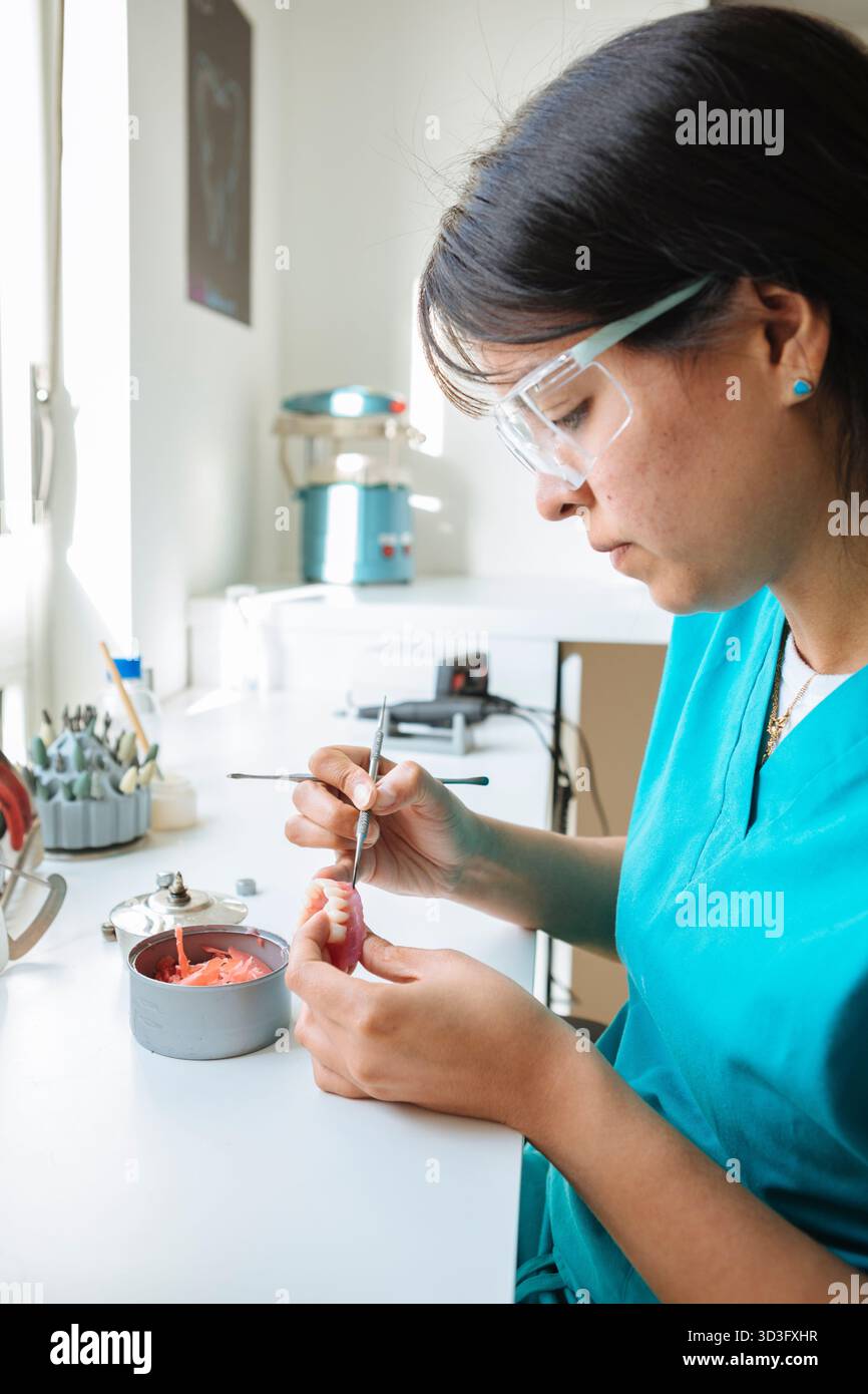 Die Seitenansicht einer fokussierten Zahntechnikerin in blauen Peelings fokussiert sich intensiv während der Herstellung einer Zahnprothese mit speziellen Werkzeugen an ihrer Arbeit Stockfoto