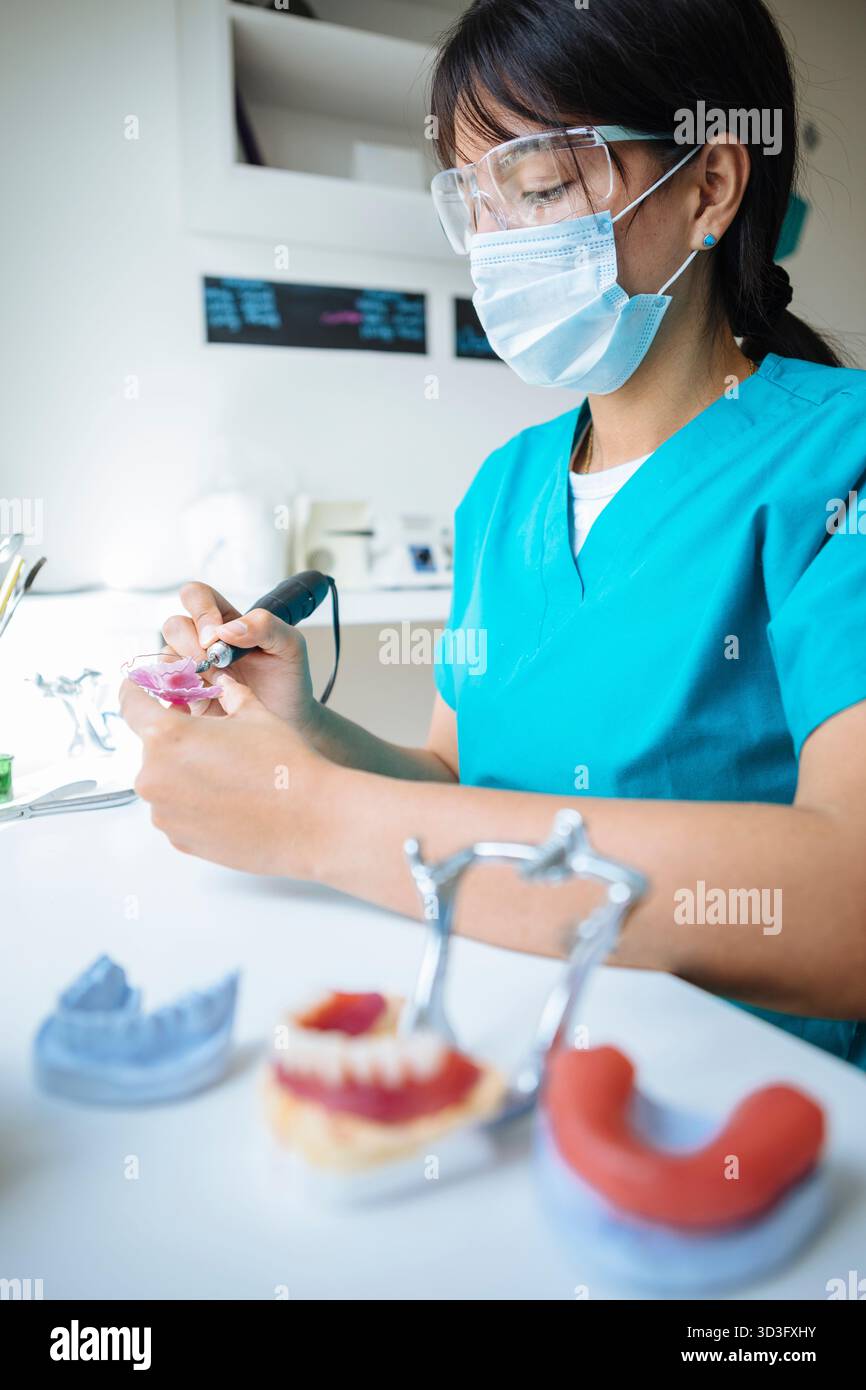 Eine Zahntechnikerin in blauen Schrubben und Schutzausrüstung fertigt an ihrem Arbeitsplatz, Showcasin, sorgfältig eine Zahnprothese mit Präzisionswerkzeugen Stockfoto