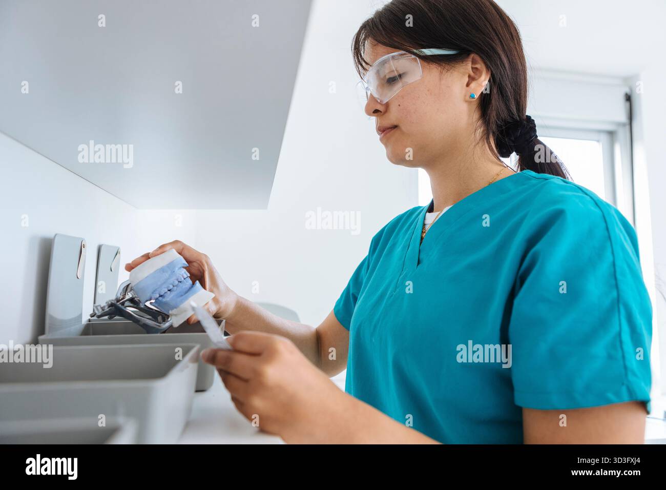 Eine Zahntechnikerin untersucht eine Zahnform genau. Sie trägt eine Schutzbrille und blaue Peelings und konzentriert sich intensiv auf ihre Arbeit in Stockfoto