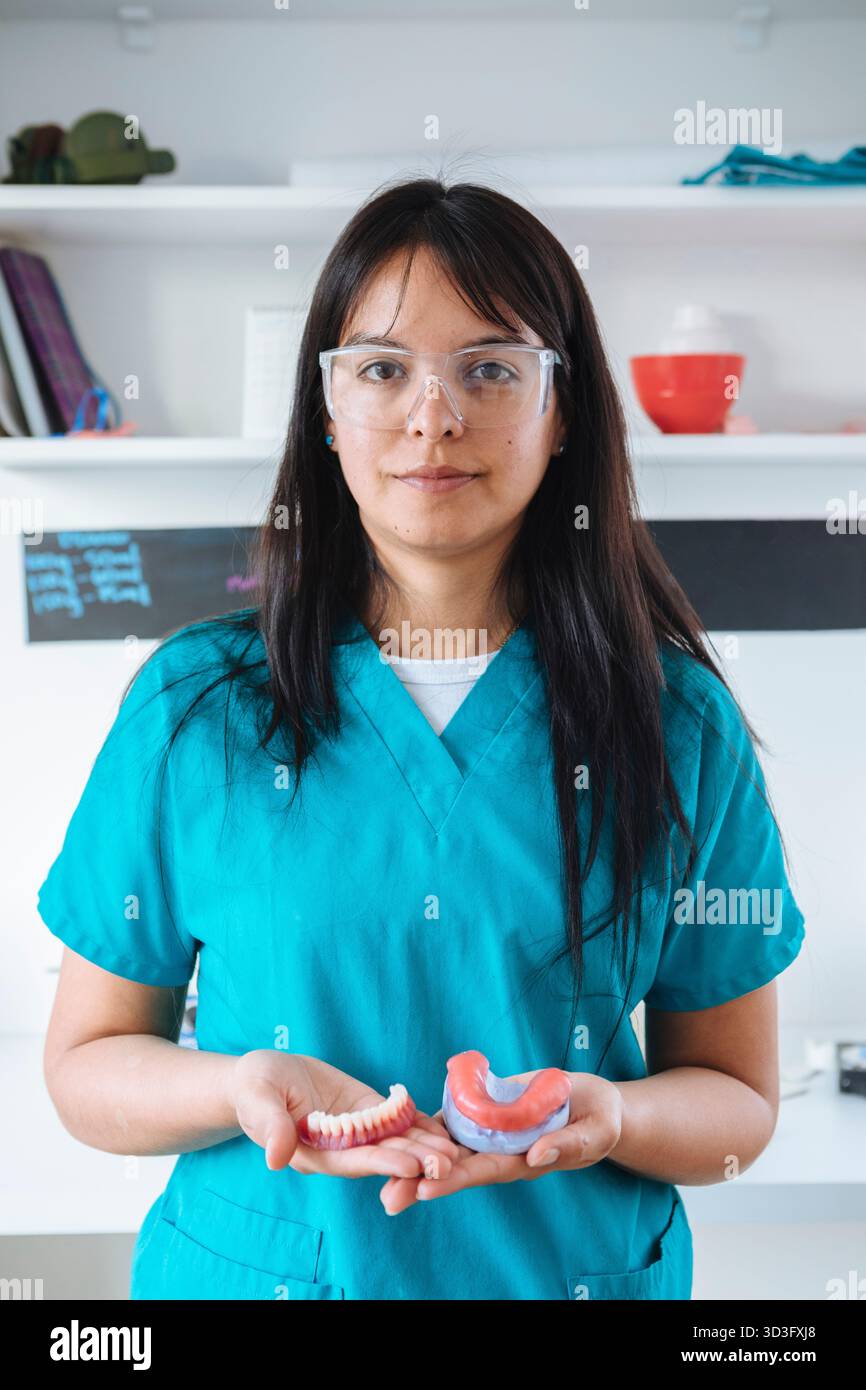 Eine Zahntechnikerin in blauen Peelings und Schutzbrille hält Zahnersatz. Sie steht selbstbewusst in einem Dentallabor und konzentriert sich auf ihr d Stockfoto