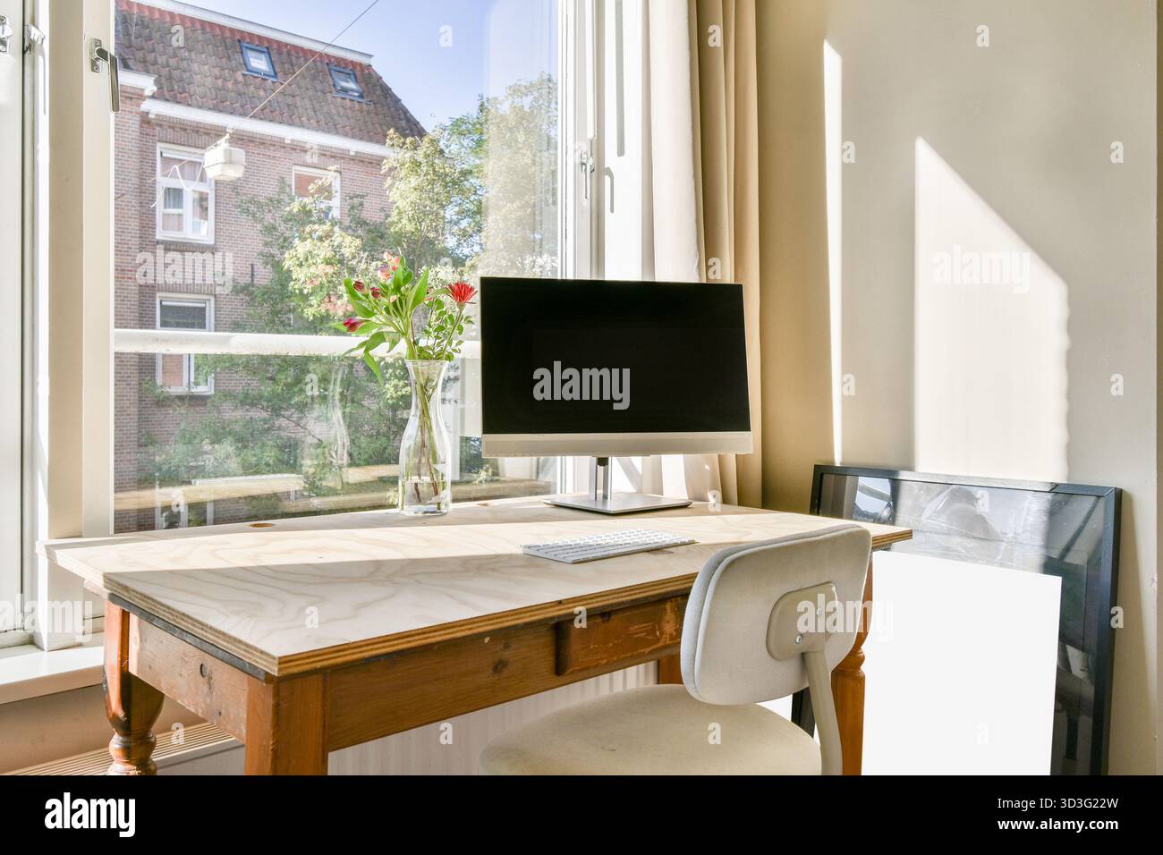 Helles Home Office mit Holzschreibtisch, Computer und minimalistischer Einrichtung. Das Sonnenlicht beleuchtet den Raum und hebt eine Vase mit Blumen und einer bezaubernden Vase hervor Stockfoto