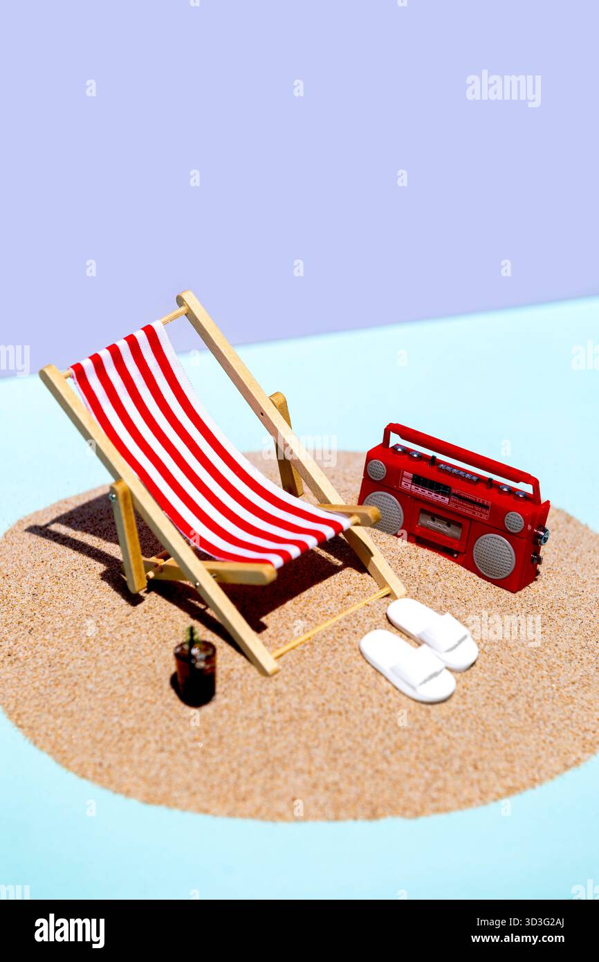 Eine verspielte Miniatur-Strandszene mit einem rot-weiß gestreiften Liegestuhl, einem leuchtend roten Radio und weißen Hausschuhen auf einem Sandkreis mit blauem bac Stockfoto