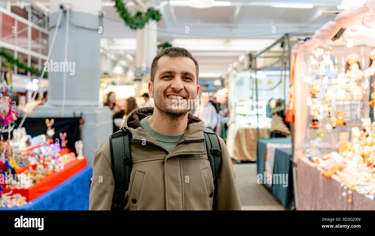 Ein glücklicher Mann mit einem Rucksack, der warm in die Kamera lächelt, während er die pulsierenden Verkaufsstände am Covent Garden Market in London erkundet und den lebhaften geldautomaten genießt Stockfoto