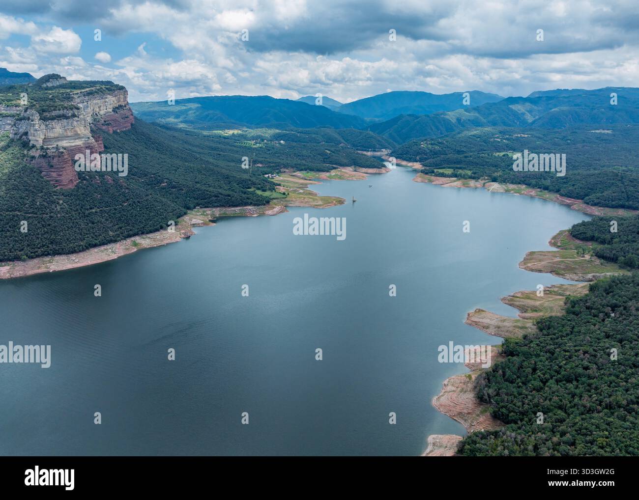 Aus der Vogelperspektive auf den ruhigen Stausee Pantà de Sau, der den Himmel spiegelt, umgeben von grünen Wäldern und zerklüfteten Klippen unter einem bewölkten Himmel, Catalunya, prov Stockfoto
