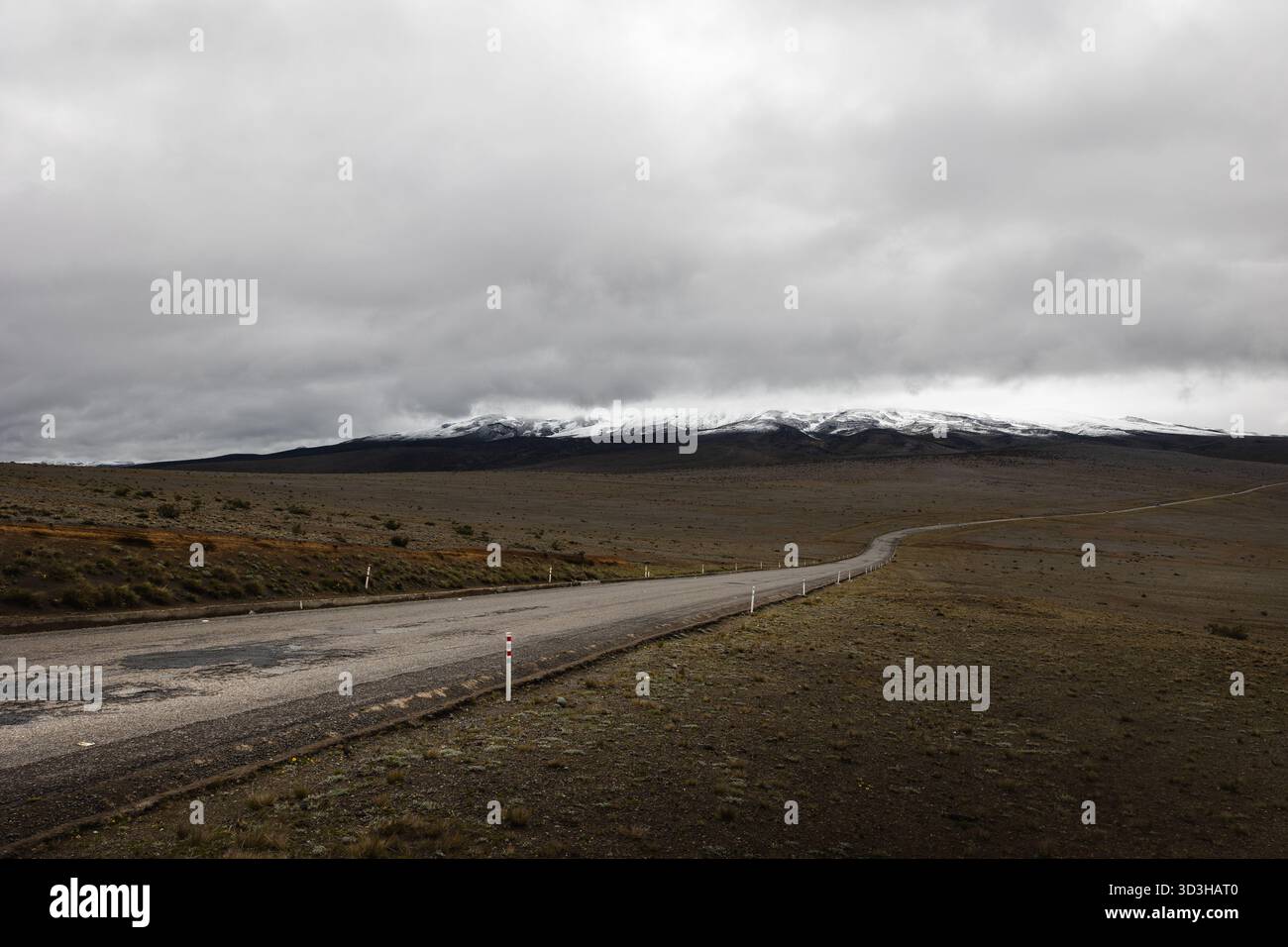 Einsame Straße, die sich unter stürmischem Himmel durch den hohen páramo schlängelt und zu den schneebedeckten Hängen des Monte Chimborazo in Ecuador führt. Stockfoto