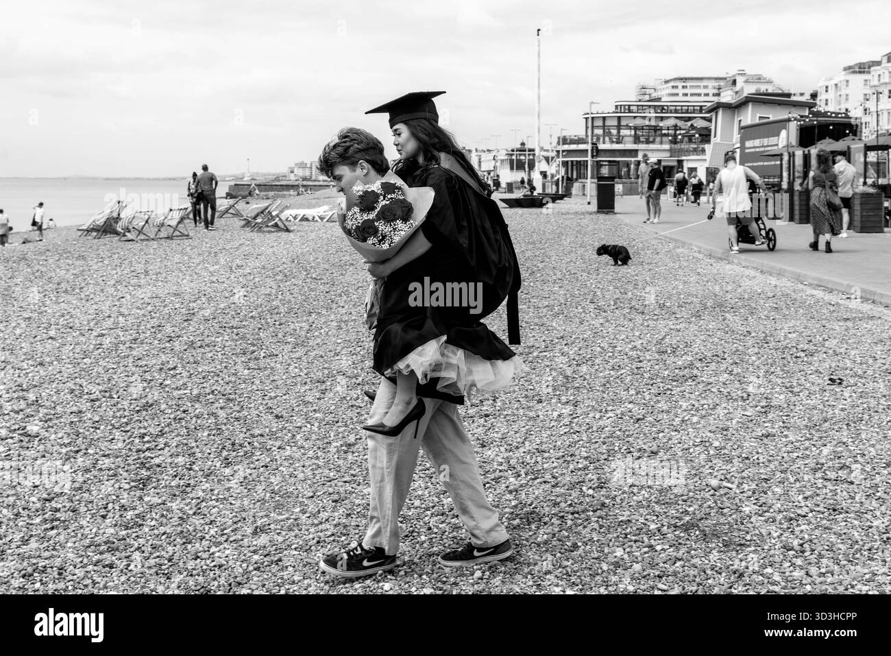 Ein junger Mann trägt seine Freundin am Strand für einige Familienfotos nach der Abschlussfeier der Universität Sussex in Brighton, East Sussex, Großbritannien. Stockfoto