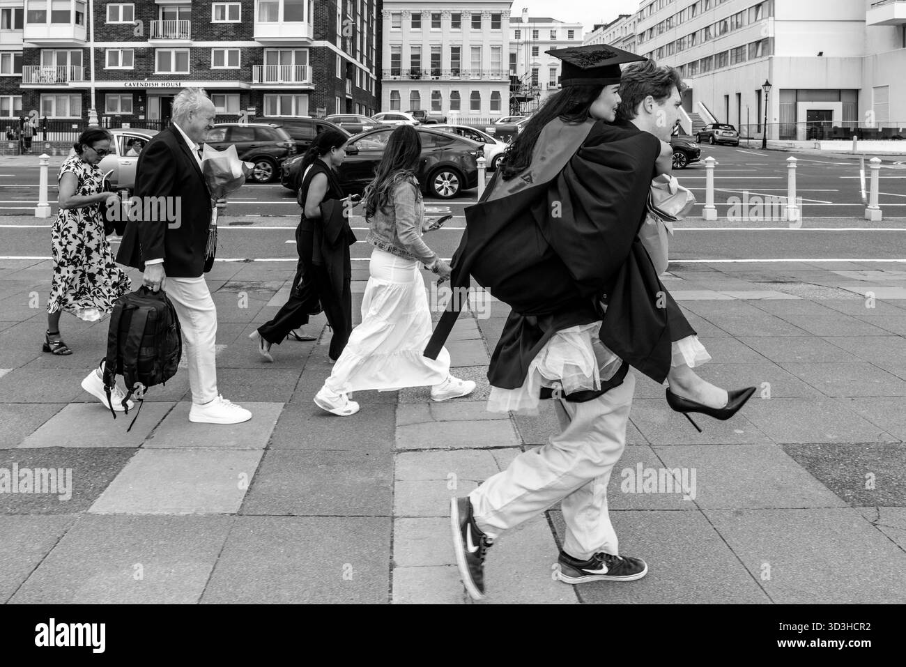 Ein junger Mann trägt seine Freundin am Meer entlang, nachdem sie sich beschwert, dass ihre Füße weh tun, University of Sussex Graduation Day, Bri Stockfoto