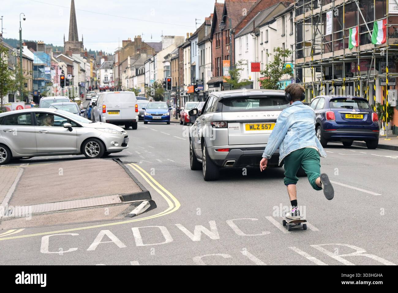 Monmouth, Monmouthshire, Wales - 23. August 2025: Person, die auf einem Skateboard hinter einem Auto auf der Hauptstraße der Stadt Monmouth fährt Stockfoto
