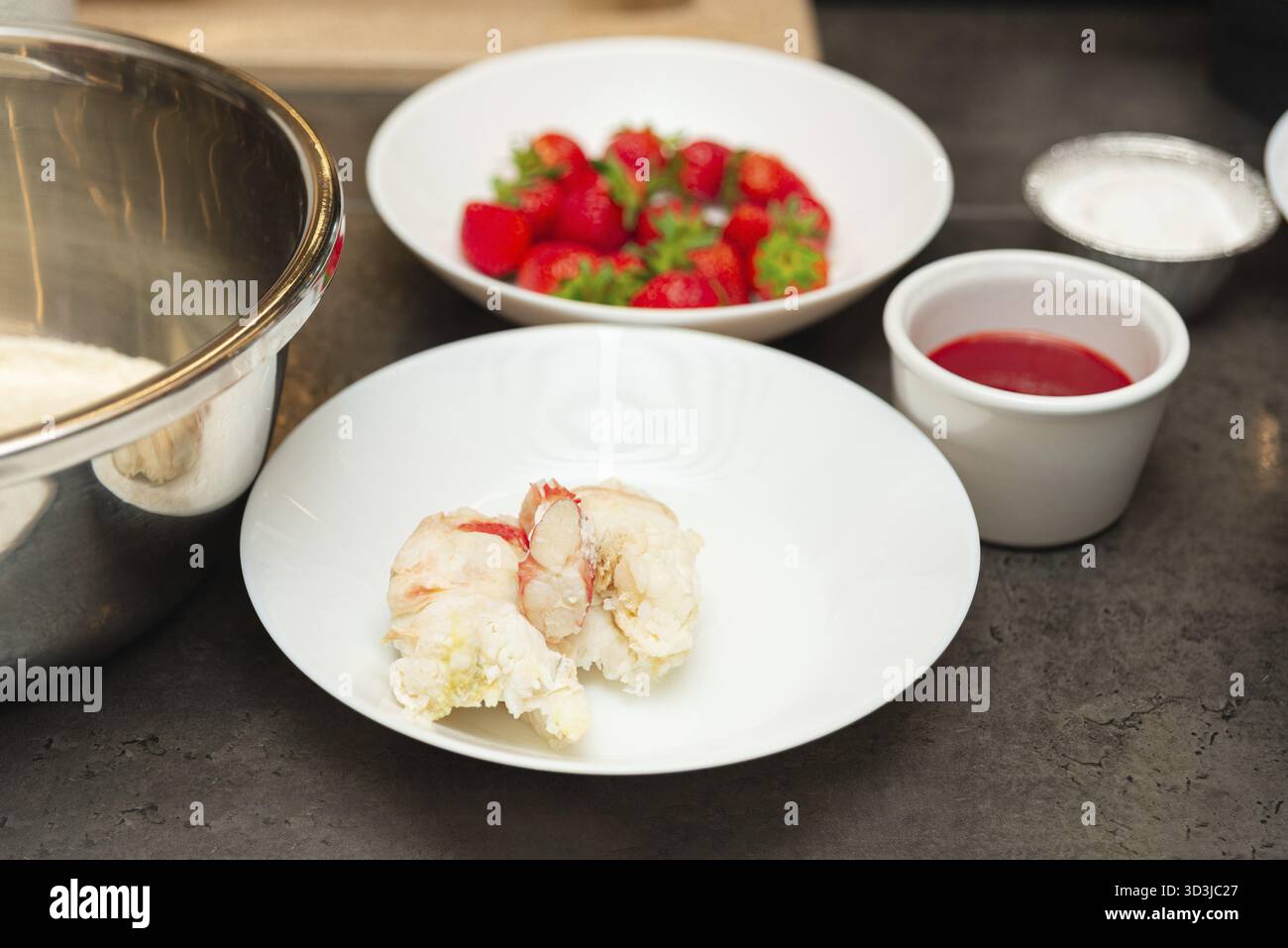 Essen, Nahaufnahme von gekochtem Hummerschwanzfleisch auf einem weißen Teller, umgeben von Erdbeeren, Soße und Zutaten für die Zubereitung auf einer Küchenzeile Stockfoto
