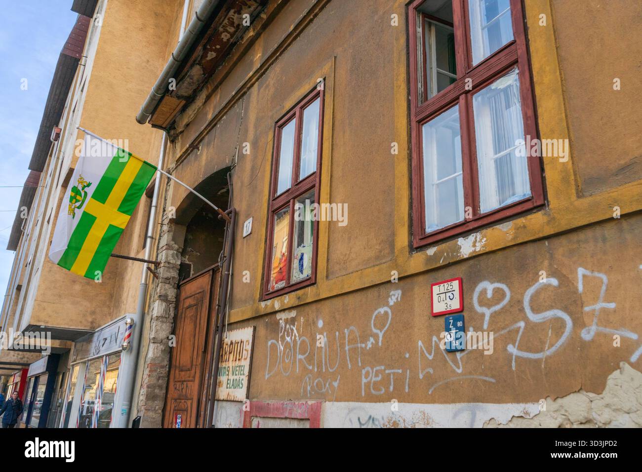 Budapester Fassade im 8. Bezirk mit Bezirksflagge, Graffiti und einem Kauf- und Verkaufsschild, das urbanen Schmutz und Straßenhandel einfängt. Stockfoto