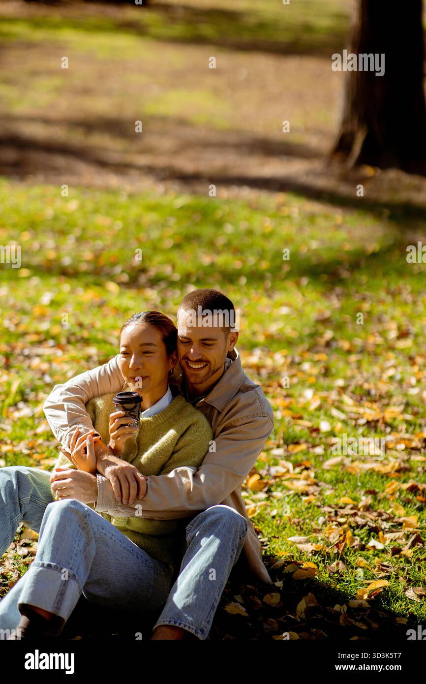 Koreanerin und Kaukasier. Ein Paar entspannt sich in einem Park voller Herbstlaub, lacht und genießt warme Getränke an einem sonnigen Nachmittag. Stockfoto
